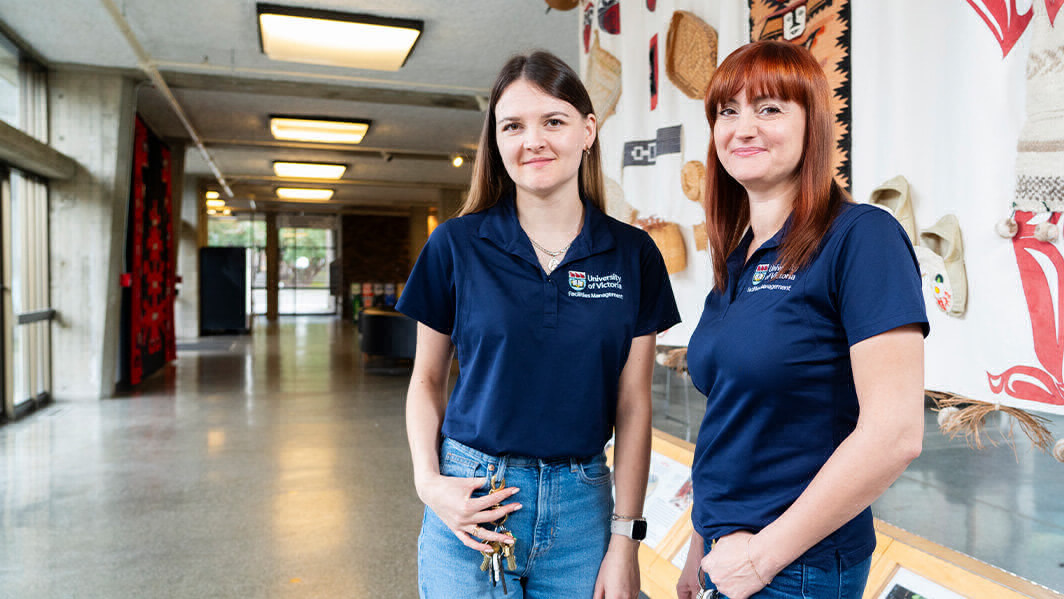Two young women wearing UVic branded golf shirts stand in a building corridor. 