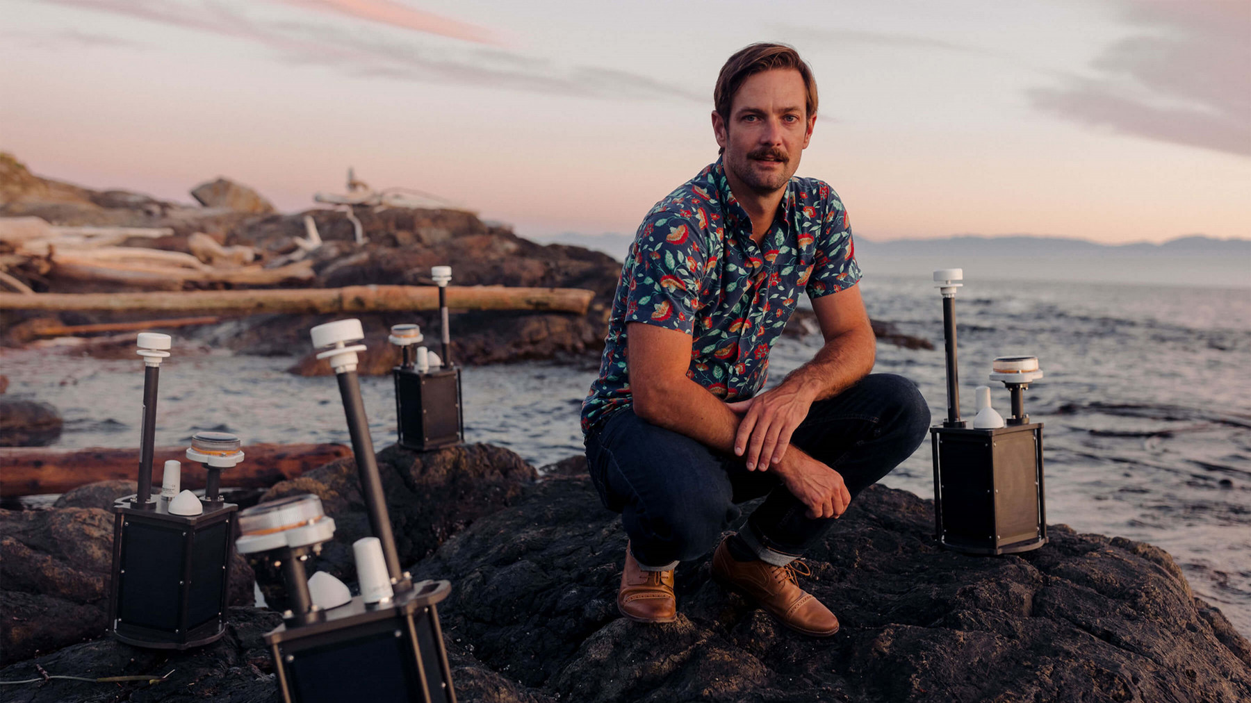 Man with a moustache and colourful short sleeve shirt crouching on a shoreline amongst electronic buoys. 