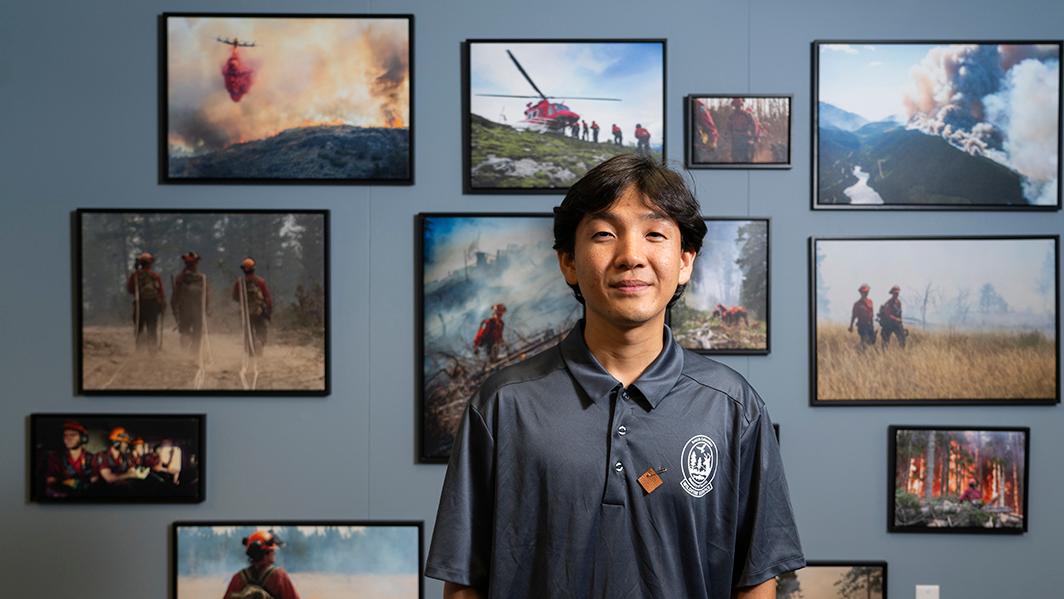 Jay Choi, a Humanities co-op student, is posing in front of a wall covered in photos of BC Wildfire Services fighting wildfires. Jay is wearing a gray collared uniform shirt and a moose hide pin.