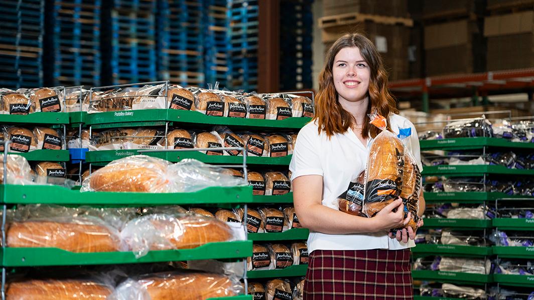Melanie is posing in a packing warehouse at Portofino Bakery, wearing a white blouse and red tartan skirt. She has long, red wavy hair. Melanie is holding up two packaged loaves of Portofino bread.