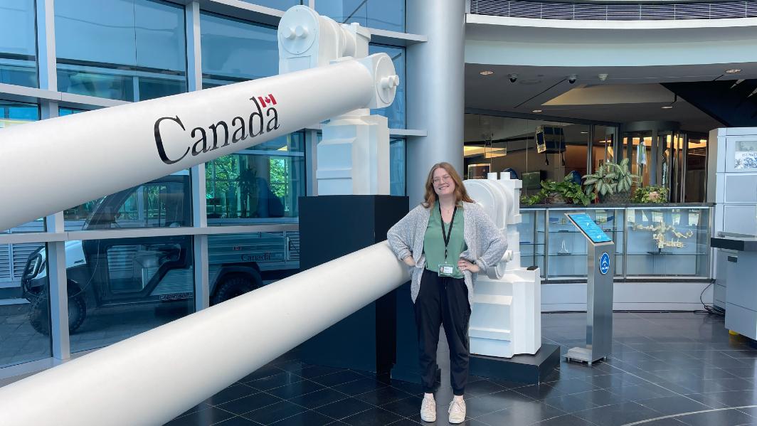 Tess Grindlay is posing in a large lab building in front of machinery emblazoned with the word "Canada."