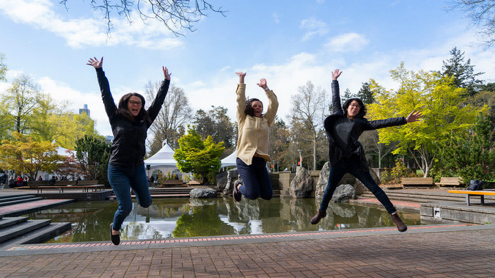 Three women jump with their hands up in front of the Petch fountain.